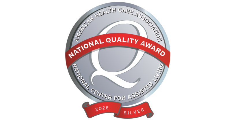 A circular badge with a silver background and black border, featuring a large stylized letter 